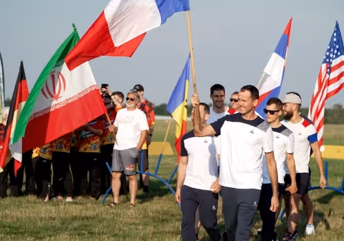 Eine Gruppe von Männern geht über eine Wiese, einige tragen die Flaggen von Frankreich, Deutschland, Iran, der Ukraine, Serbien und den USA.