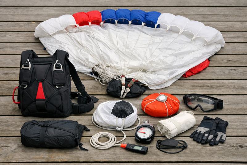 Complete professional skydiving gear laid out on a wooden floor – main parachute, reserve parachute, harness, altimeter and skydiving goggles.