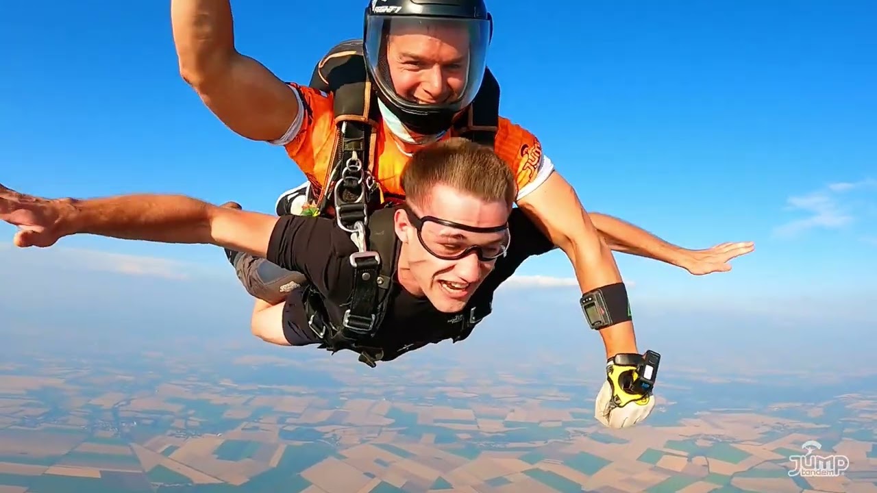 Náhled videa ze dvou kamer: pasažér v černém tričku a tandem pilot v oranžovém ve volném pádu nad poli